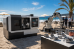 June 2026 AI manufacturing releases displayed as CNC machine tools, Physical AI robots and machined parts in a sunny beach launch scene